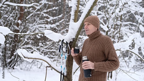 XC skier dressed in tweed sweater and knit hat makes a break in winter forest and drinks a cup of coffee from a thermos