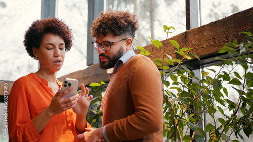 Two colleagues standing in modern office, actively discussing project using smartphone