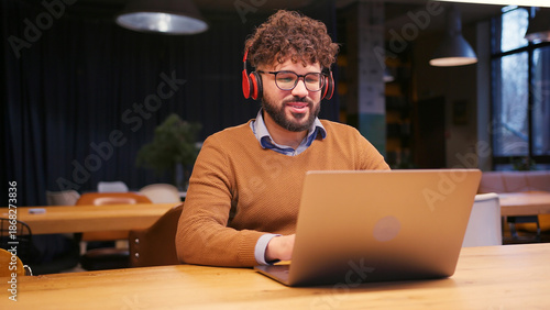 Slika na platnu Middle eastern businessman wearing stylish eyeglasses in headphones, using lapto
