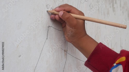 a small boy's hand is drawing or scribbling on a white wall using a simple wooden pencil