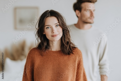 Portrait of young Caucasian couple indoors. Woman in foreground, looking directly at camera. Man blurred in background, looking away. Focus on connection and modern lifestyle