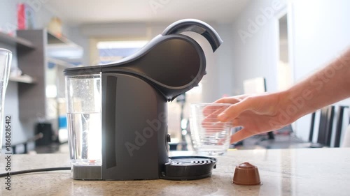 Coffee capsule machine preparing a drink as a hand places a glass cup on the drip tray in a modern kitchen or office.