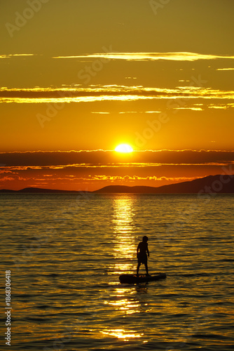 Silhouette of young boy on SUP stand up paddle board at shiny golden sunset