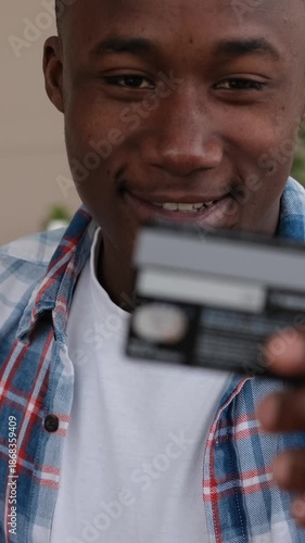 A smiling young African American man holds up a credit card, ready to make a payment or shop online. Vertical video.