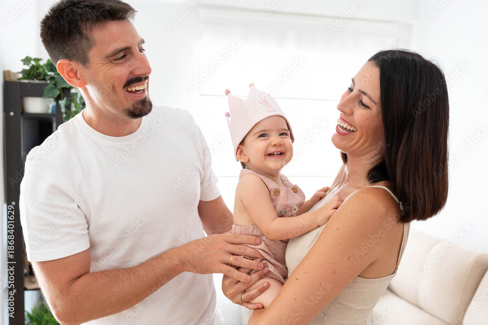 Fototapeta premium Happy family couple celebrating baby's first birthday at home, parents holding and laughing with their adorable one year old daughter wearing a crown, enjoying togetherness and love