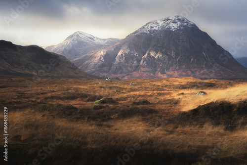 Dramatic golden light on the dark, moody, mountain landscape of Buachaille Etive Mor with a distant Blackrock Cottage at Glencoe in the Scottish Highlands, Scotland.