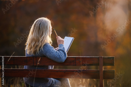 Bilde på lerret Thoughtful woman sitting on park bench and writing diary or inspiration notes during sunset