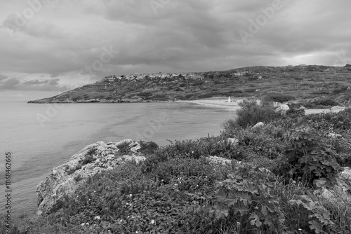 Black and white coastal landscape with calm Mediterranean sea, sandy shoreline and rolling hills, creating a timeless and minimalist scene with dramatic clouds and a quiet, contemplative atmosphere.