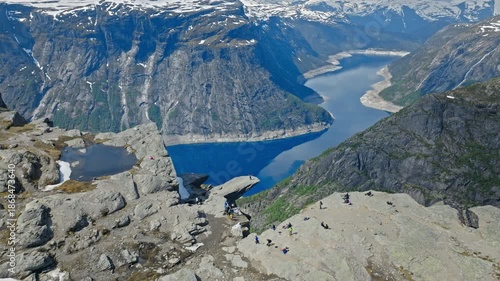 Rocky mountain slope descending toward the fjord at Trolltunga. Aerial summer view combining rugged stones, snow traces, and vivid blue water below.