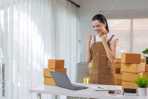 Asian woman feeling happy and successful, celebrating a new order or good sales from her online business, packing products at home