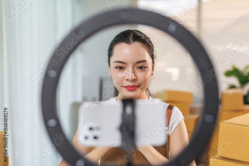 Young asian woman creating content for her online audience using a smartphone, ring light, and cardboard boxes