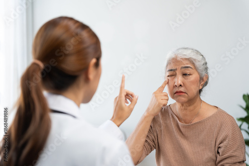 Asian doctor examining senior woman's eye, pointing a finger for eye movement test. Patient feels discomfort or pain