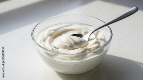 A brightly lit close-up shot captures fresh white, thick greek yogurt being scooped with a metal spoon from a clear glass bowl on a simple white tabletop.