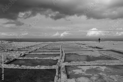 Traditional salt pans along the rocky coastline show geometric stone basins and coastal textures, captured in black and white at Saline, Malta, on 29 December 2025.