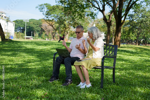 Happy senior Asian couple celebrating success while using a laptop on a bench in a sunny public park.