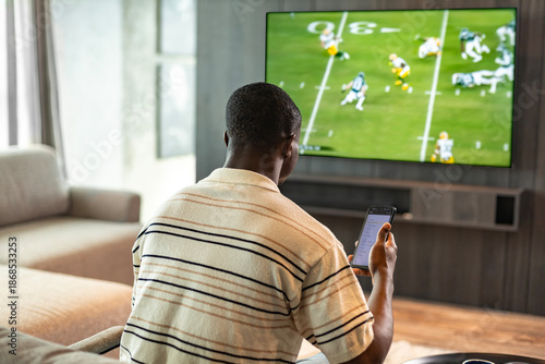 Obraz na płótnie African man holding smartphone with sports bets app while watching American football match at home in living room