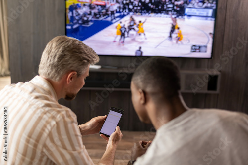 Foto Two friends holding smartphone with sports bets app while watching basketball match at home in living room