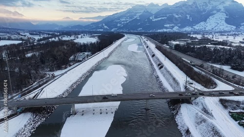 Drone flying over a snow-covered sandbank and bridge on the Rhine River between Liechtenstein and Switzerland, showing cars crossing and winter river scenery.