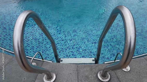 Footage of Clean swimming pool ladder with stainless steel handrails and blue tiled water.Calm ripples create a relaxing background for luxury hotel, resort, spa, wellness, summer travel, and leisure.