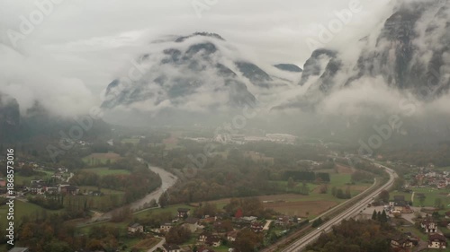 Scenic view of Italian mountain valley with river and highway infrastructure. Small village houses scattered among autumn trees under low clouds and heavy morning mist in Alpine region.