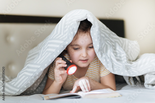 A thoughtful young girl lies on a cozy bed, deeply engrossed in reading an open book under the covers with flashlight