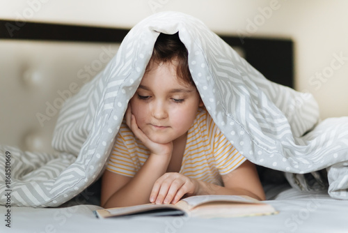 A thoughtful young girl lies on a cozy bed, deeply engrossed in reading an open book