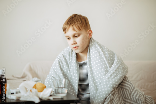 A young sick kid boy sits on a bed, looking thoughtful and unwell.