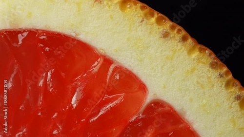 Macro close up of a red grapefruit slice slowly rotating, highlighting vibrant citrus flesh, rind texture, and glossy details against a black background. Ideal for food, color, and abstract concepts.