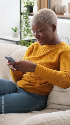 Vertical video of a smiling young African American woman relaxing on a couch at home and texting on her mobile phone.