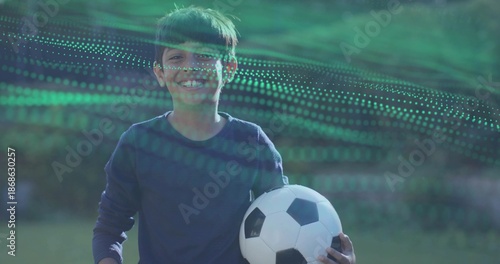 Smiling Indian-Asian boy holding soccer ball on sports field, wearing navy shirt with green overlay