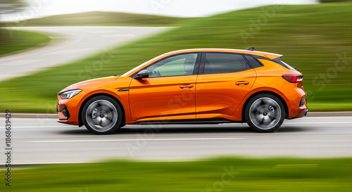 Vibrant orange modern hatchback dynamically driving at speed along an asphalt road with blurred green countryside scenery creating motion