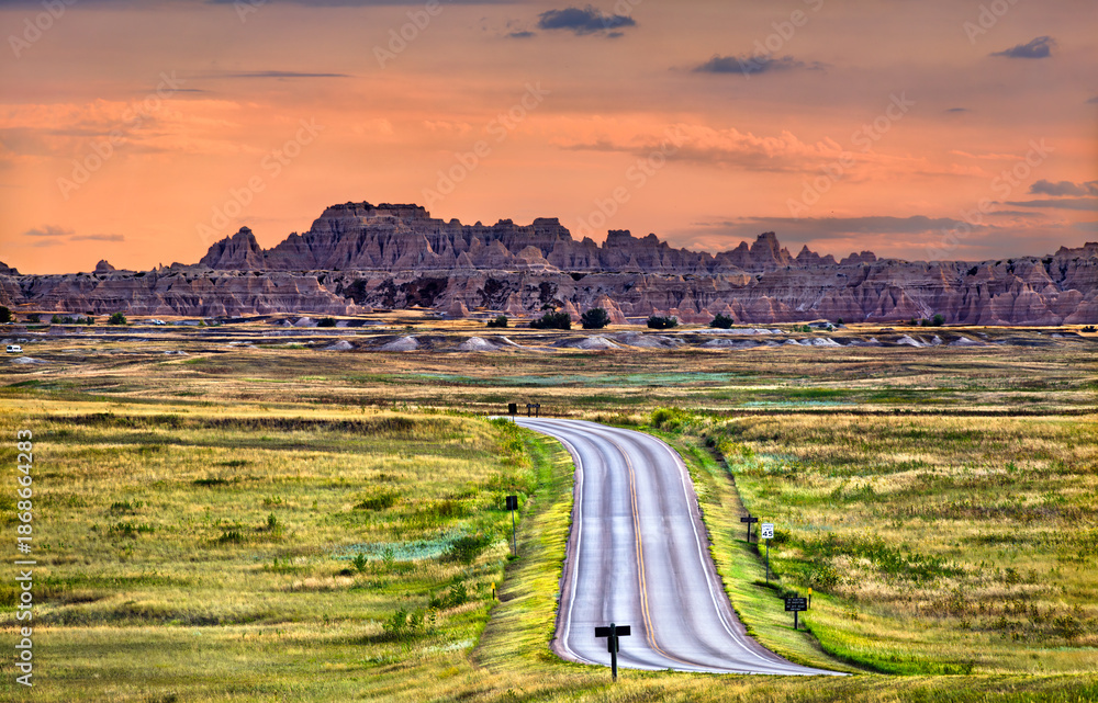 Fototapeta premium Badlands National Park landscape stands in South Dakota, USA. Scenic view features the winding Loop Road leading towards rugged rock formations and green prairie under a colorful sunset sky