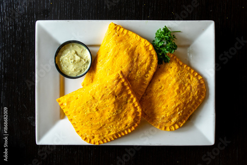 Overhead view of golden baked Jamaican beef patties on a white plate with cilantro sauce on a black table