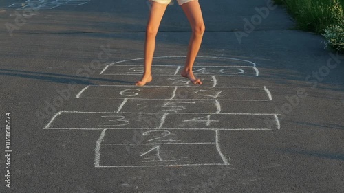 A child stands barefoot on a hopscotch drawing on the pavement. The sun shines bright, casting shadows on the ground in a neighborhood