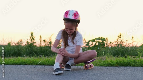 A girl in a helmet sits on the ground after falling off her skateboard, holding her knee. The sun is setting in the background, casting warm light over the scene