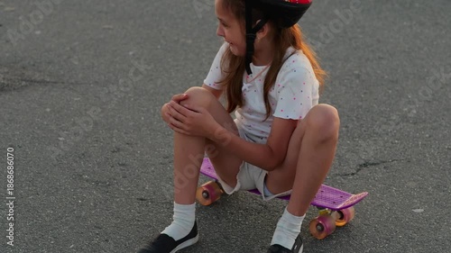 A young girl enjoys her time sitting on a skateboard on the pavement. She wears a helmet and seems happy while playing outside in the late afternoon light