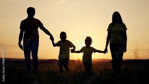 Family walking hand in hand across open field at sunset silhouette of parent mother father and child holding hand together in tall grass under warm orange sky slow stroll toward horizon with glow