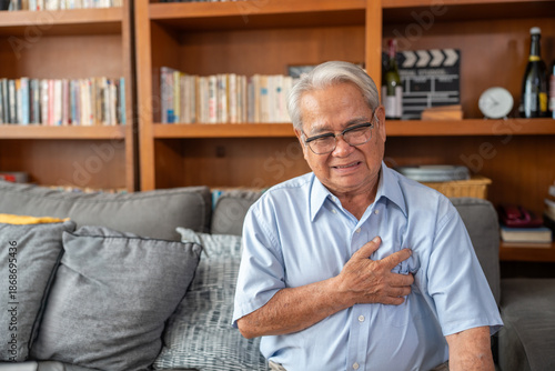 Senior man experiencing chest pain at home. Older man sitting on couch with hand on chest. Heart disease problems.