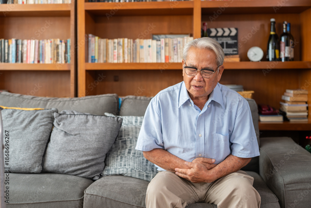 Fototapeta premium Asian elderly man holding stomach pain while sitting on the sofa at home. Senior man experiencing digestive issues or abdominal cramps.