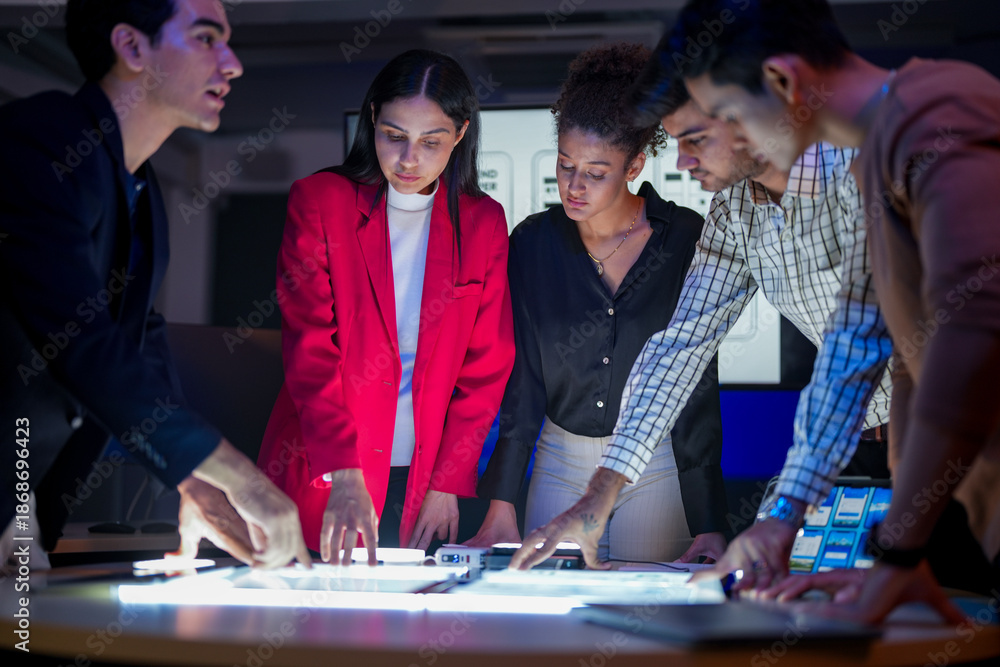Fototapeta premium Creative UI/UX design team analyzing mobile app wireframes on illuminated digital touch table. Diverse developers brainstorming web layout strategy in modern high-tech office at night.