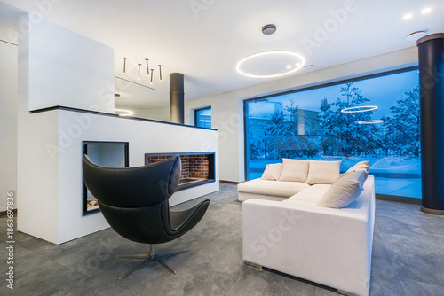 An incomparable view of snow-covered fir trees from the window of a luxurious living room. Twilight. A fireplace, a white sofa and a chic armchair in the interior.