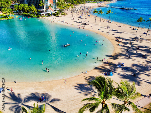 Aerial view of the Hilton Lagoon's turquoise waters meet the sandy beach, dotted with palm trees and sunbathers under a bright sky, Honolulu, Hawaii, United States.