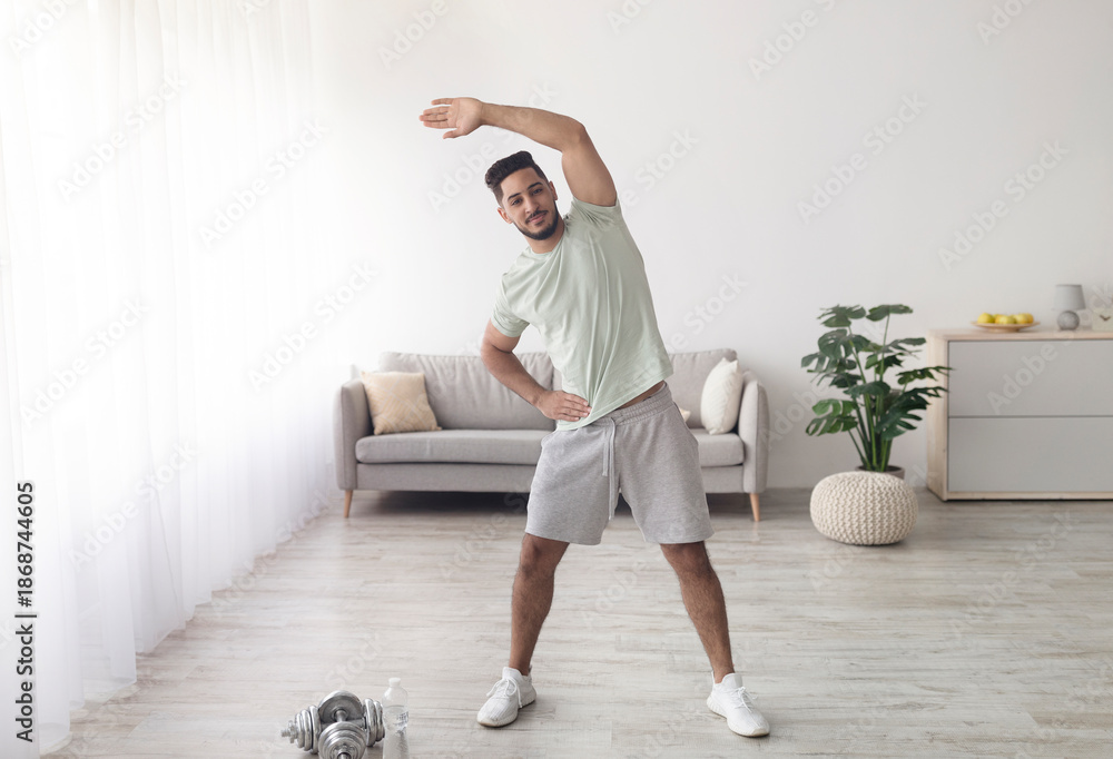 Fototapeta premium Sporty young Arab man exercising at home during coronavirus lockdown, full length portrait. Attractive middle Eastern guy working out on domestic training, copy space