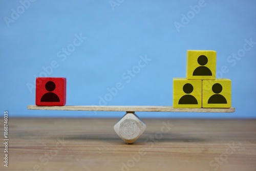 Wooden balance scale shows a single red block on one side and a stacked group of yellow blocks on the other, illustrating teamwork, structure, and varied roles in business.