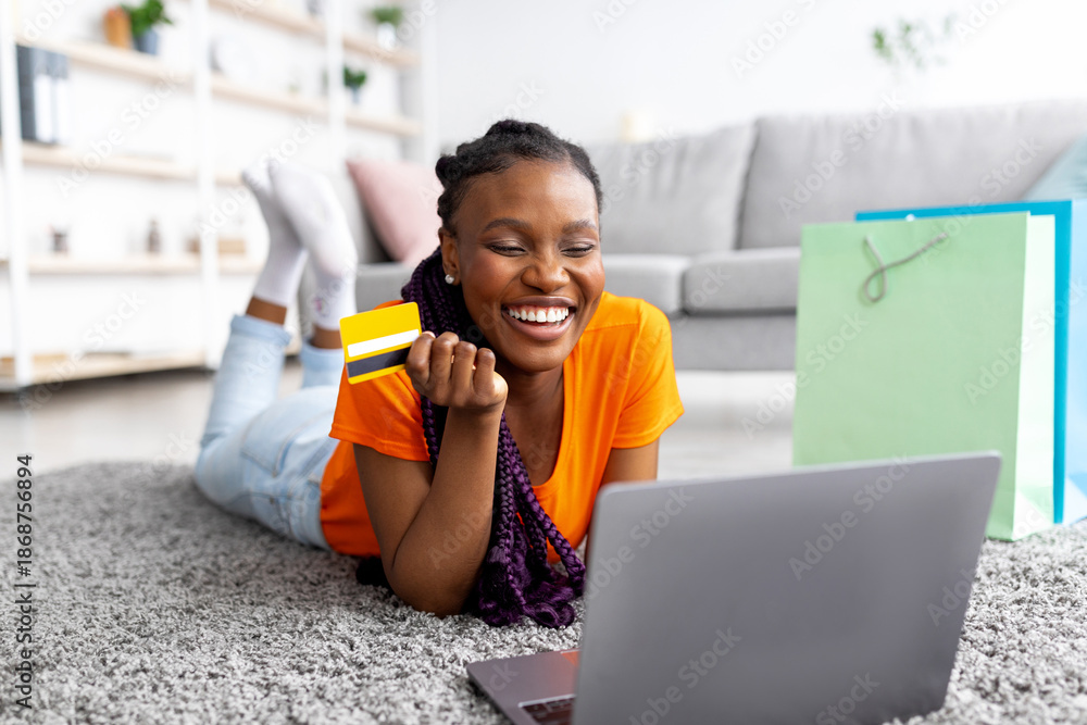 Φωτογραφία Young African American woman buying online with credit card and laptop, lying on floor at home