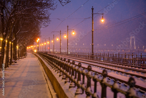 Budapest, Hungary - January 6, 2026: Snow-covered tram tracks stretch along the Danube embankment in Budapest at night, lined by glowing vintage streetlamps under a purple-hued winter sky.
