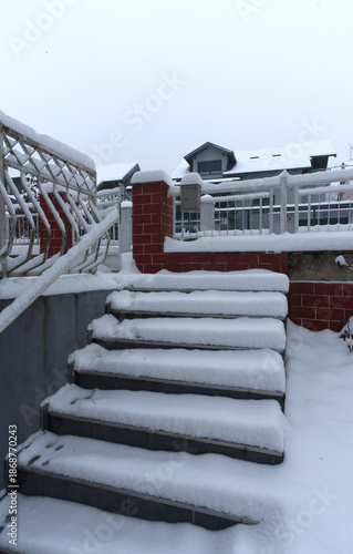 A snow-covered outdoor staircase leads through a quiet residential area, framed by metal railings and brick fencing. Fresh snowfall blankets the steps and rooftops, evoking a serene winter atmosphere.