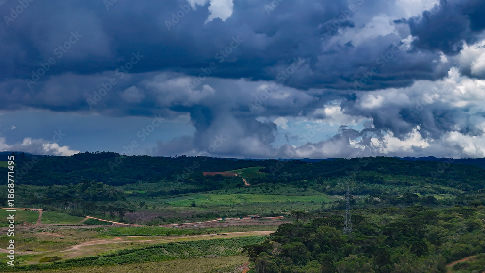 Obraz premium The image shows a landscape of lush green hills and forests under a dramatic, cloud-laden sky, with areas of open land and what appears to be a column of rain in the distance.