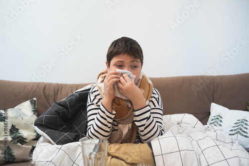 Sick child sitting on sofa wrapped in blanket, holding tissue to nose, winter illness concept, home interior, cold or flu symptoms.