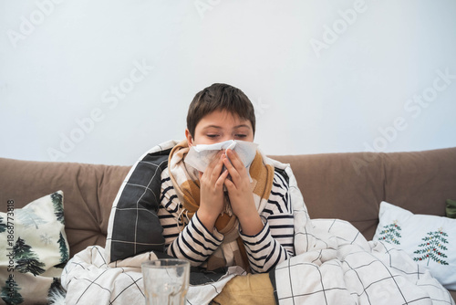 Sick child sitting on sofa wrapped in blanket, holding tissue to nose, winter illness concept, home interior, cold or flu symptoms.